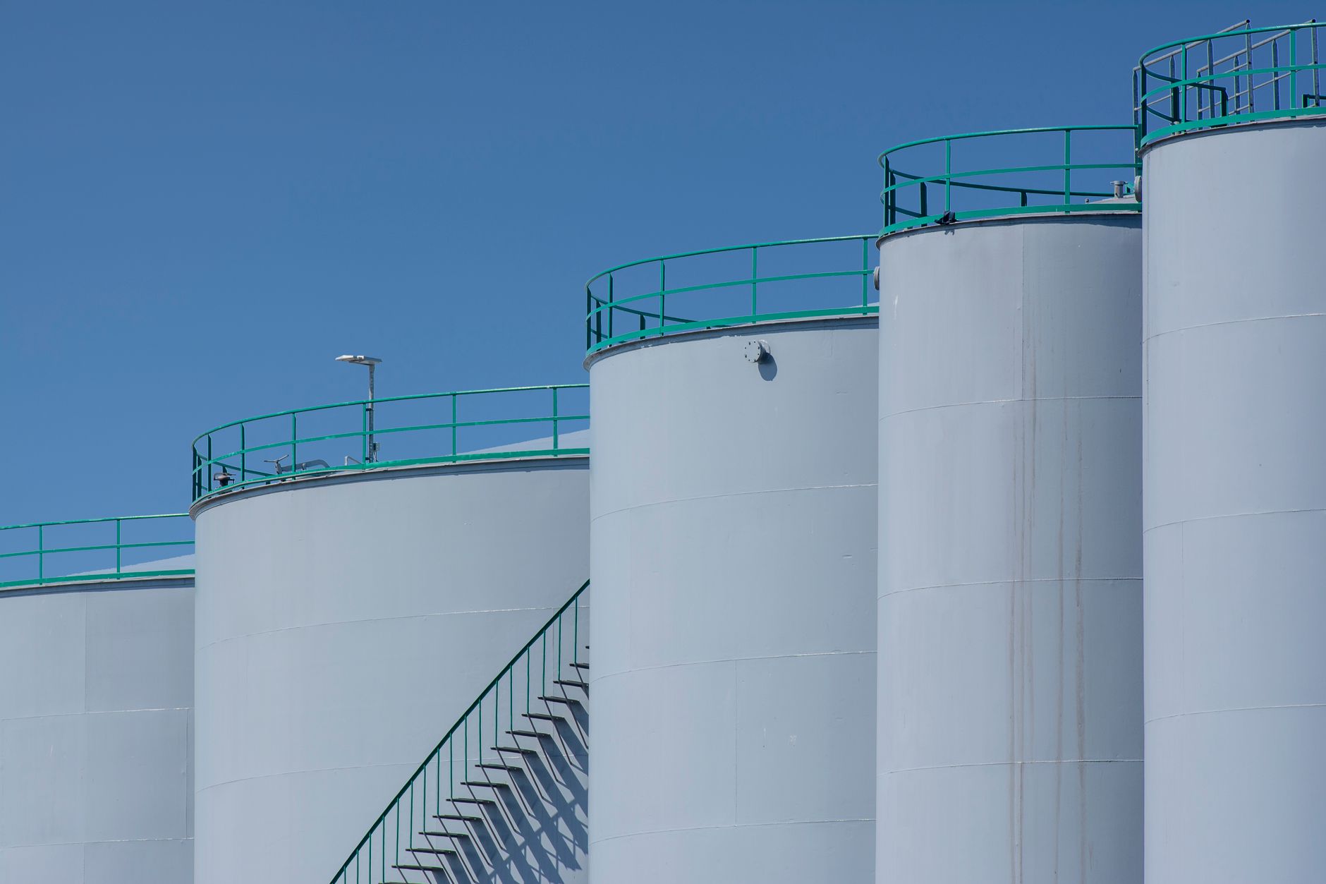 Row of white crude storage tanks with green handrails against a clear sky