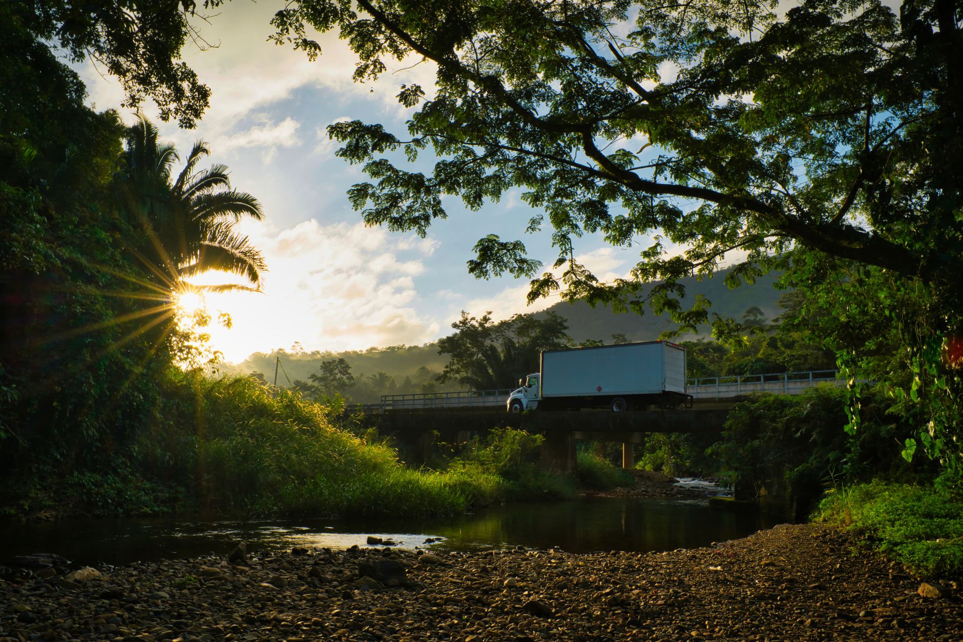 Box truck crossing a low concrete river bridge at sunrise